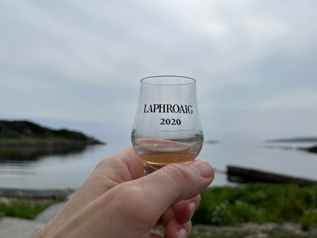 Hand holding glass of Laphroaig whisky by a bay