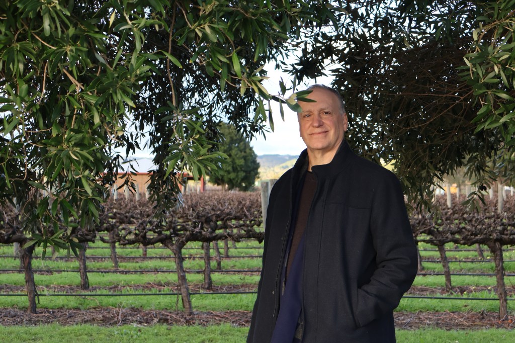 Man (Doctor D) standing in front of vines and olive trees