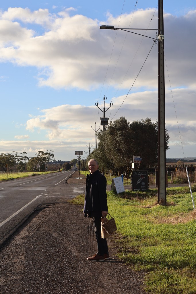 Man standing by country roadside with basket and brown paper bag on a sunny day

